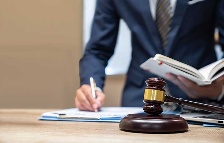 Gavel in image forefront, man in suit writing on a notepad and holding a book in background